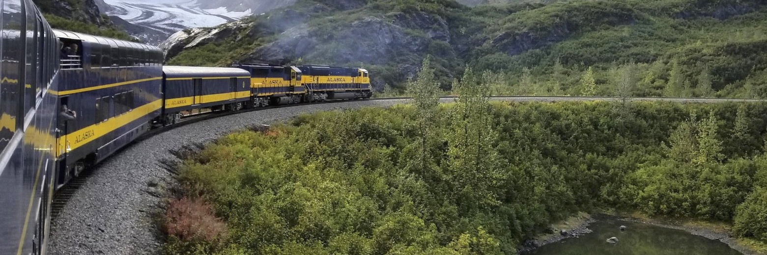 Alaska: a blue and yellow train curving along tracks past a mountain glacier and a small pond